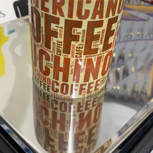 May include: A white ceramic coffee mug with a brown and orange design featuring the words "Americano", "Espresso", "Cappuccino", "Latte", and "Coffee" repeated in a pattern.