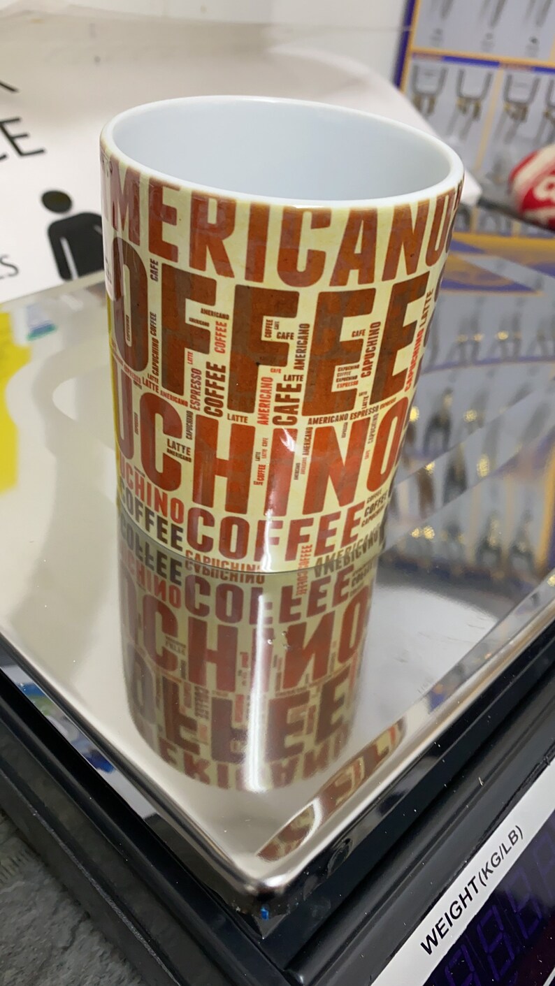 May include: A white ceramic coffee mug with a brown and orange design featuring the words "Americano", "Espresso", "Cappuccino", "Latte", and "Coffee" repeated in a pattern.