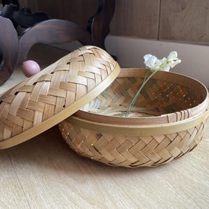 May include: A round, woven bamboo basket with a matching lid, partially open. The basket is light brown with a woven pattern. A sprig of white flowers rests inside. The lid is off to the side.