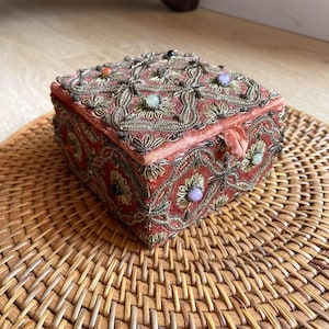 May include: A small, square, coral-colored box with intricate gold embroidery and colorful gemstone accents. The box is made of a soft, velvety material and features a hinged lid. The box is sitting on a woven, circular mat.