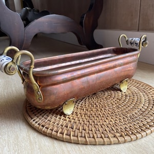 May include: A copper planter with brass handles and feet, resting on a woven mat. The planter features decorative blue and white ceramic handles. The copper has a rich, aged patina, adding to its vintage appeal. A decorative piece for home decor.