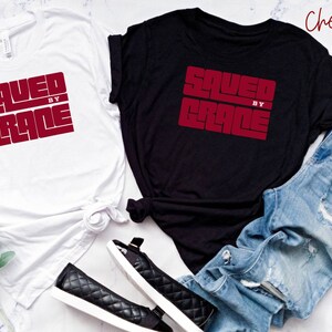 May include: Two t-shirts with the text "SAVED BY GRACE" in red lettering on a white and black background. The shirts are laid out on a white surface with a pair of black shoes and blue jeans.