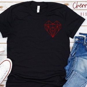 May include: Black t-shirt with a red geometric heart design. The shirt is laid flat on a white wooden surface with a pair of white sneakers and blue jeans.