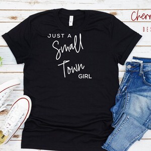 May include: Black t-shirt with white text that reads "Just a Small Town Girl". The shirt is laid flat on a white wood surface with a pair of white sneakers and blue jeans.