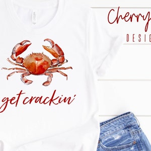 May include: White t-shirt with a watercolour crab design in red and brown. The words "get crackin'" are written in red script below the crab. The words "Cherry Vixen DESIGNS" are written in red script at the top right.