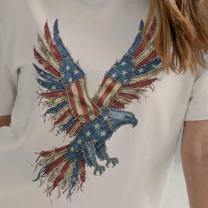 Può includere: T-shirt color crema con una grafica dettagliata di un'aquila calva americana con le ali spiegate. Le ali dell'aquila sono disegnate con le stelle e le strisce della bandiera americana. Il corpo dell'aquila è blu e la maglietta ha maniche corte.