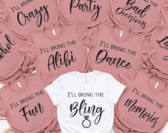 May include: A selection of pink t-shirts with black text, each bearing the phrase "I'll Bring The" followed by words such as "Crazy", "Party", "Bad Decisions", "Alcohol", "Alibi", "Dance", "Laughs", "Fun", "Memories", and "Bling". One white shirt says "I'll Bring The Bling".