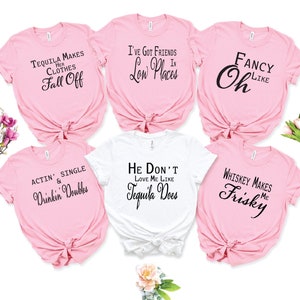 May include: Six pink and white t-shirts with funny sayings about alcohol. The sayings include "Tequila Makes Her Clothes Fall Off", "I've Got Friends In Low Places", "Fancy Oh Like", "Actin' Single & Drinkin' Doubles", "He Don't Love Me Like Tequila Does", and "Whiskey Makes Me Frisky".