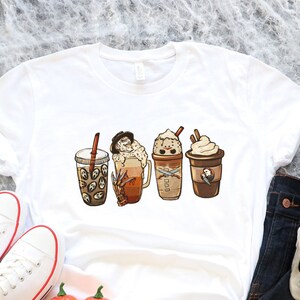 May include: White t-shirt featuring a graphic of four different coffee drinks. The drinks are decorated with autumnal elements such as pumpkins, leaves, and candy corn. The t-shirt is laid out on a black surface with a skeleton hand, jeans, and pumpkins.