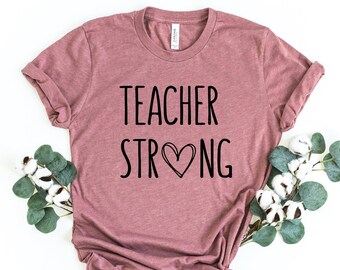 May include: A dusty rose-colored t-shirt with the words "TEACHER STRONG" in black script, with a heart symbol replacing the letter "O". The shirt is styled with a knot at the bottom and decorated with sprigs of greenery and white flowers.