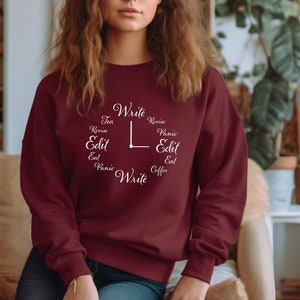 May include: A maroon sweatshirt with a clock design featuring writing-related words like "Write," "Edit," and "Revise." The clock hands point to the time. The sweatshirt is a solid color and has long sleeves.