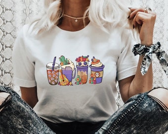 Könnte beinhalten: Ein weißes T-Shirt mit einem Aufdruck von fünf bunten Bechern mit Totenköpfen und Blumen. Die Person, die das Shirt trägt, sitzt mit gekreuzten Beinen und trägt zerrissene Jeans und Leoparden-Sneaker.