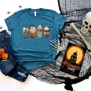 May include: Teal blue t-shirt featuring a graphic of four Halloween-themed coffee drinks. The t-shirt is lying on a black mesh fabric with a skull and crossbones, and a Halloween party invitation with the text "Halloween Party 31st October 2020".
