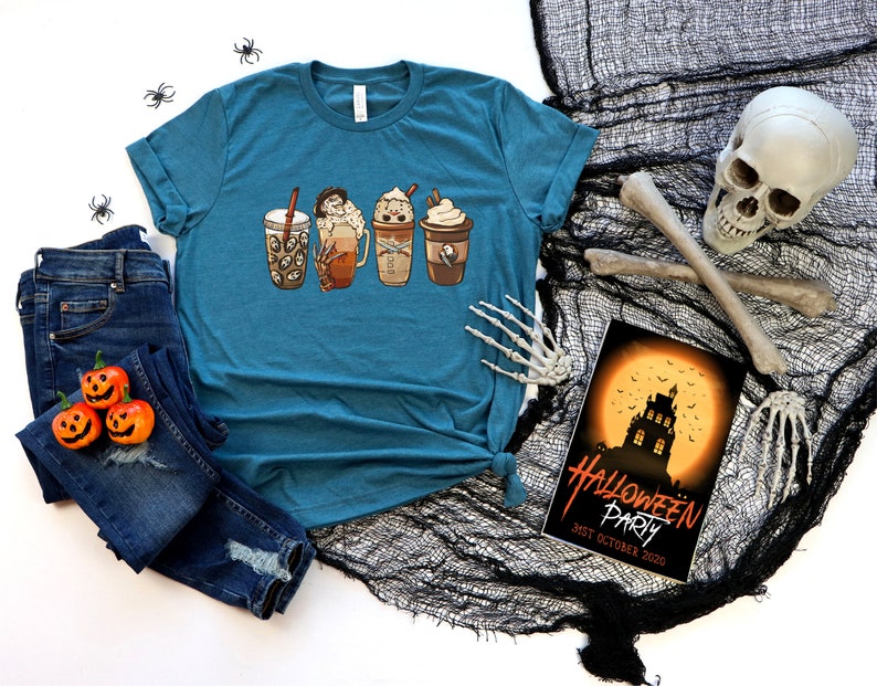 May include: Teal blue t-shirt featuring a graphic of four Halloween-themed coffee drinks. The t-shirt is lying on a black mesh fabric with a skull and crossbones, and a Halloween party invitation with the text "Halloween Party 31st October 2020".