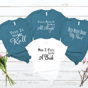 May include: Four t-shirts with different sayings on them. The shirts are teal blue and white. The sayings are "This is how we roll", "A little messed up, but we're all alright", "Man I feel like a bride", and "Beer never broke my heart".