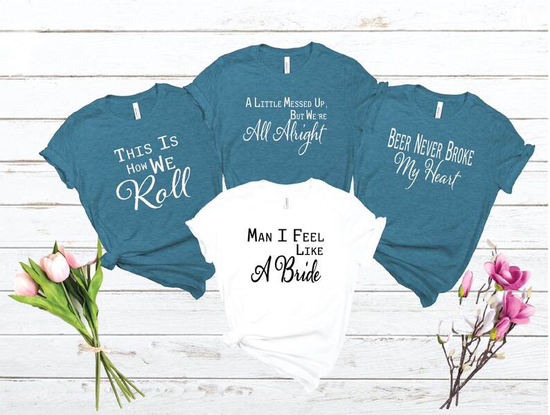 May include: Four t-shirts with different sayings on them. The shirts are teal blue and white. The sayings are "This is how we roll", "A little messed up, but we're all alright", "Man I feel like a bride", and "Beer never broke my heart".