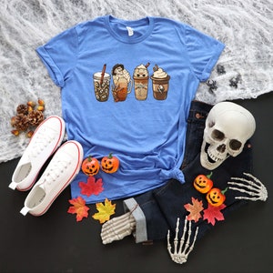 May include: A blue t-shirt featuring a graphic of four different coffee drinks. The drinks are all in different cups and have various toppings. The shirt is displayed on a table with a skull, jeans, and autumn leaves.