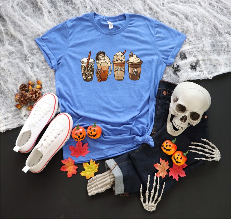 May include: A blue t-shirt featuring a graphic of four different coffee drinks. The drinks are all in different cups and have various toppings. The shirt is displayed on a table with a skull, jeans, and autumn leaves.