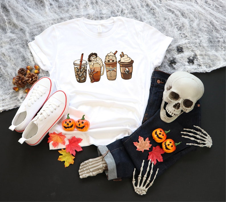 May include: White t-shirt featuring a graphic of four different coffee drinks. The drinks are decorated with autumnal elements such as pumpkins, leaves, and candy corn. The t-shirt is laid out on a black surface with a skeleton hand, jeans, and pumpkins.