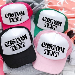 May include: Four trucker hats in pink, black, green, and pink. Each hat has a white front panel with the words "CUSTOM TEXT" in black. The hats are on a light-colored surface with sunglasses and a towel.