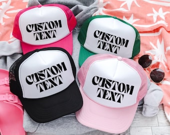 May include: Four trucker hats in pink, black, green, and pink. Each hat has a white front panel with the words "CUSTOM TEXT" in black. The hats are on a light-colored surface with sunglasses and a towel.