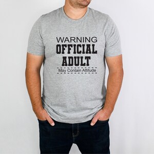 May include: A gray t-shirt with the text "WARNING OFFICIAL ADULT May Contain Attitude" printed in black.