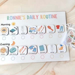 以下が含まれることがあります： 「RONNIE'S DAILY ROUTINE」のテキストが入った子供向けの毎日のルーチンチャート。チャートには、「起きる」、「朝食を食べる」、「就寝時間」など、朝と夕方のイラスト付きのタスクが含まれています。追加のタスクカードの小さなバッグも含まれています。
