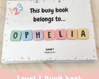 Peut inclure: Un livre d'activités de niveau 1 blanc avec le texte "Ce cahier d'activités appartient à..." et le nom "OPHELIA" épelé avec des tuiles de lettres colorées. Le livre a une reliure à spirale et est décoré de confettis colorés.