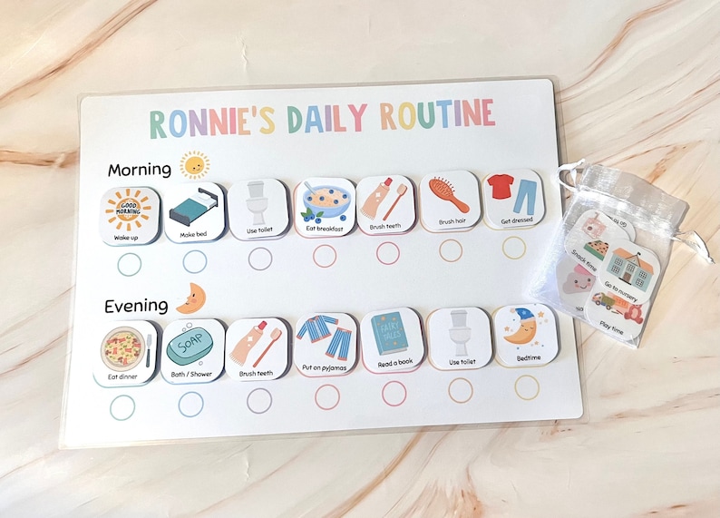 以下が含まれることがあります： 「RONNIE'S DAILY ROUTINE」のテキストが入った子供向けの毎日のルーチンチャート。チャートには、「起きる」、「朝食を食べる」、「就寝時間」など、朝と夕方のイラスト付きのタスクが含まれています。追加のタスクカードの小さなバッグも含まれています。