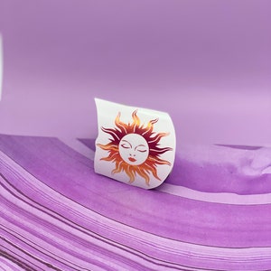 May include: A white sticker with a gold and orange sun with a closed eye face design.