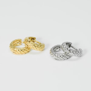 Peut inclure: Deux paires de créoles texturées. Une paire est de couleur or et l'autre de couleur argent. Les boucles d'oreilles présentent un motif répétitif de petites bosses en relief sur leur surface. Les boucles d'oreilles sont présentées sur un fond blanc.