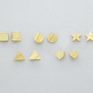 Puede incluir: Una colección de pendientes de botón dorados en varias formas: cuadrados, círculos, estrellas, triángulos y corazones. Cada pendiente tiene una superficie texturizada con un aspecto de metal cepillado. Los pendientes están colocados sobre un fondo gris claro.