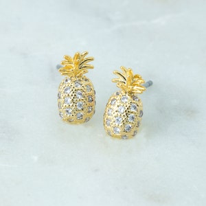 Op de afbeelding: Een paar goudkleurige ananas oorknopjes. De oorbellen hebben een textuuroppervlak met kleine, heldere kristallen. De ananas toppen zijn gedetailleerd met een textuur, stekelig ontwerp. De oorbellen worden gepresenteerd op een wit marmeren oppervlak.