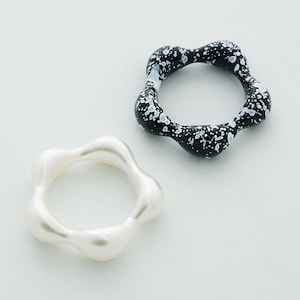 May include: Two rings with a unique, organic shape. One ring is a glossy black with white speckles, and the other is a smooth, pearly white. The rings have a wavy, flower-like design.