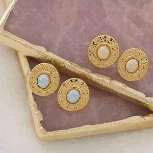 May include: Four gold-colored stud earrings with an oval, opalescent center stone. Each earring is surrounded by small, clear crystals and a textured gold border. The earrings are displayed on a pink and gold surface.