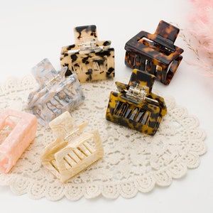 May include: Six square hair claw clips in various colors and patterns. The clips include shades of pink, cream, tortoiseshell, and marbled blue and white. They are arranged on a white lace doily, with a pink floral accent in the background.