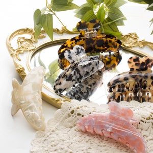 May include: Four hair clips in a variety of colors and patterns, including tortoise shell, black and white, and pink. The clips are arranged on a gold tray with a mirror and a white lace doily.