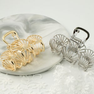 May include: Two metal hair claw clips, one gold and one silver, each featuring a seashell design. The clips are displayed on a marble-patterned surface with a white lace trim, showcasing a beach-inspired aesthetic.