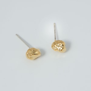 May include: A pair of gold-colored stud earrings. Each earring features a textured, irregular shape, resembling a small nugget. The posts are silver-colored. The earrings are displayed against a white background.