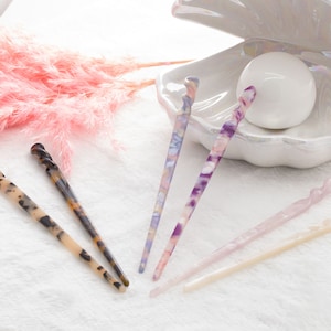 May include: A collection of hair sticks in various colors and patterns. The sticks are made of a marbled material in shades of brown, purple, and pink. They are arranged on a white surface with a decorative seashell and pink feathery accents.