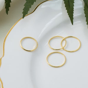 Puede incluir: Cuatro anillos finos de color dorado se colocan sobre un plato blanco con borde dorado. Los anillos tienen un diseño sencillo y pulido. Se ven hojas verdes en el fondo.