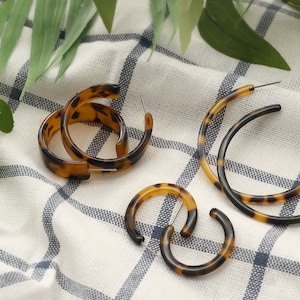 May include: Several pairs of hoop earrings in various sizes. The earrings have a tortoiseshell pattern in shades of brown and black. They are displayed on a white and blue checkered cloth, with green leaves in the background.