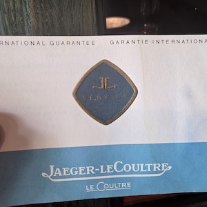 May include: A white and blue Jaeger-LeCoultre International Guarantee card. The card features the Jaeger-LeCoultre logo in a blue diamond shape. The text "INTERNATIONAL GUARANTEE" and "JAEGER-LECOULTRE" are visible.