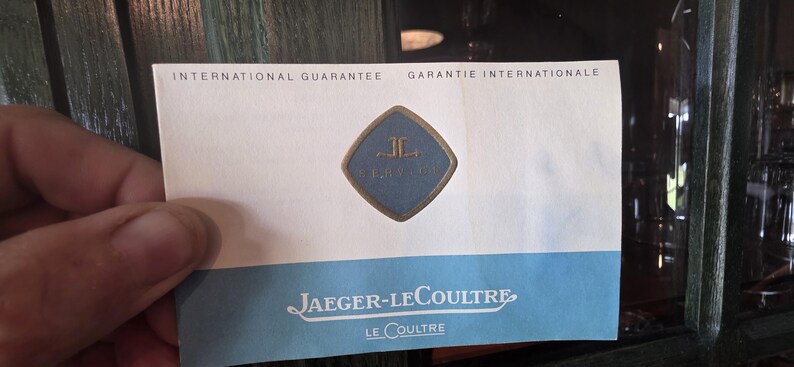 May include: A white and blue Jaeger-LeCoultre International Guarantee card. The card features the Jaeger-LeCoultre logo in a blue diamond shape. The text "INTERNATIONAL GUARANTEE" and "JAEGER-LECOULTRE" are visible.