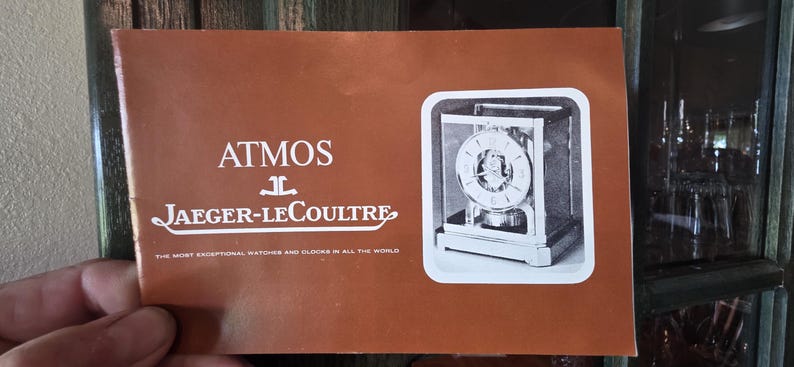 May include: A brochure with the text "ATMOS JAEGER-LECOULTRE" in white on a brown background. The brochure features a black and white image of an Atmos clock. The text at the bottom reads "THE MOST EXCEPTIONAL WATCHES AND CLOCKS IN ALL THE WORLD."