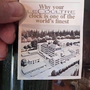 May include: A vintage advertisement for LeCoultre clocks, held by a hand. The ad features an illustration of a building complex and text that reads "Why your LeCoultre clock is one of the world's finest."