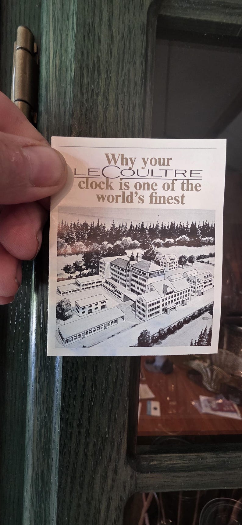 May include: A vintage advertisement for LeCoultre clocks, held by a hand. The ad features an illustration of a building complex and text that reads "Why your LeCoultre clock is one of the world's finest."