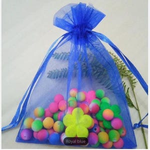 Puede incluir: Una bolsa de regalo de organza azul real llena de cuentas de colores y un dije de flor amarilla. La bolsa tiene un cierre de cordón y está hecha de tela transparente.