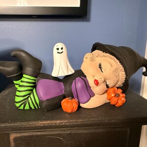 May include: A ceramic figurine of a reclining character wearing a black witch hat, a purple top, and green and black striped leggings. The figure has closed eyes, red lips, and is accompanied by a small white ghost and two orange pumpkins.
