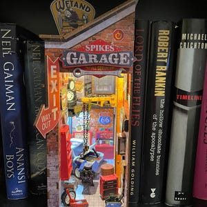 May include: A miniature garage diorama with the sign "Spikes Garage" and "Full Service". The interior features a car, tools, and a "66" sign. The diorama is placed between several books, including "Anansi Boys" and "Lord of the Flies".